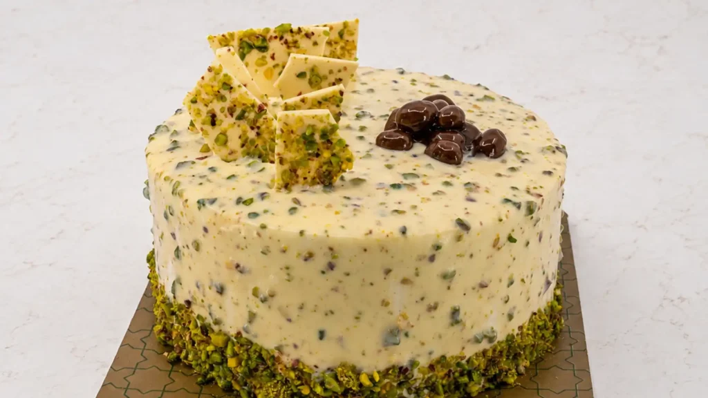 Pistachio cake with white chocolate glaze, a chopped pistachio border, pistachio bark shards and chocolate covered nuts on top
