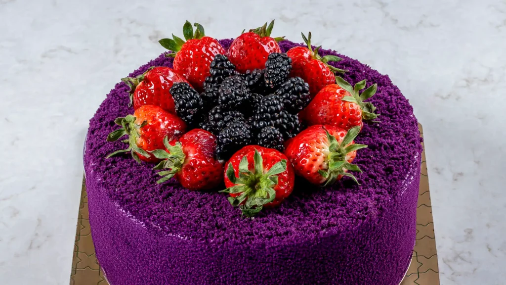 Round purple crumb coated layer cake topped with fresh strawberries and blackberries on a cake board